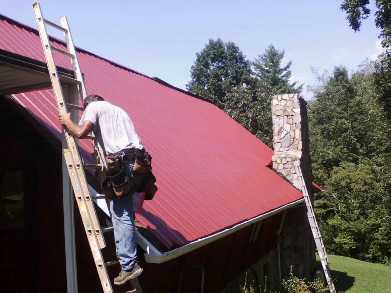 Roofer on a ladder beside a red metal roof and stone chimney — Soffit & Fascia Repair by Roofing Brothers in East Grand Forks
